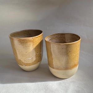 Peut inclure: Deux tasses en céramique faites à la main. Les tasses ont une forme arrondie avec une base beige clair et une partie supérieure tachetée de couleur beige. Les tasses sont probablement destinées à boire des boissons.