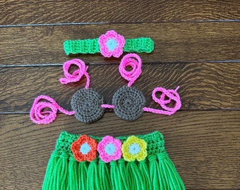 May include: A crocheted hula outfit set in bright green, brown, and pink. The set includes a headband, a bikini top, and a skirt with fringe. The headband and skirt are adorned with colorful flower accents.