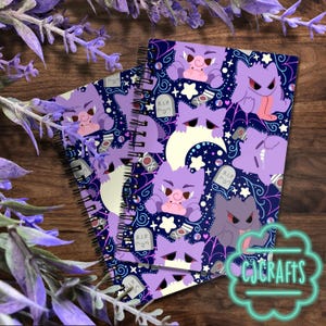 May include: Two spiral-bound notebooks with a purple and navy blue pattern featuring cartoon ghosts, tombstones, and stars. The notebooks have a dark wood background with purple floral accents and the text "CJ CRAFTS".