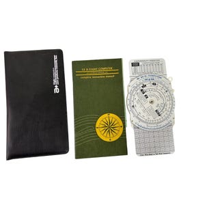 May include: A white E6-B flight computer with a green instruction manual and a black leather case. The computer has a circular slide rule with a compass rose and a gridded chart.