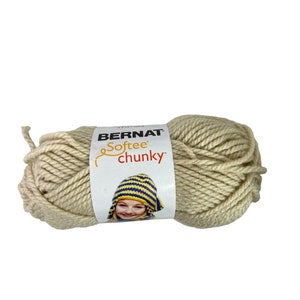 May include: A skein of Bernat Softee Chunky yarn in a cream color. The yarn is thick and textured, with a label featuring the brand name and a child wearing a blue and yellow hat.