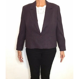 May include: A dark purple blazer with a single button closure and two front pockets. The blazer is worn over a white top and paired with black pants. The blazer's fabric has a subtle texture.