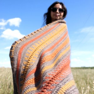 May include: A close-up of a colorful crocheted blanket with stripes of orange, brown, and yellow. The blanket is draped over a person's shoulders, with the person's face out of focus.