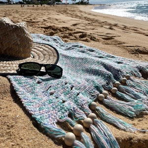 Pode incluir: Uma toalha de praia com um padrão azul, verde e rosa e contas de madeira. A toalha tem franjas na parte inferior e está sobre uma praia de areia com um chapéu de palha e óculos de sol em cima.
