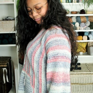 May include: A woman wearing a striped crocheted sweater with pink, white, and gray stripes. The sweater has a v-neck and a pocket on the front.