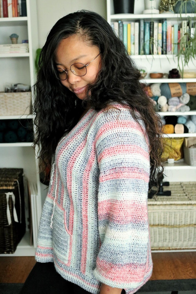 May include: A woman wearing a striped crocheted sweater with pink, white, and gray stripes. The sweater has a v-neck and a pocket on the front.