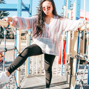 May include: A person wearing a pink and white striped sweater, black leggings, and white sneakers is standing on a playground structure. The structure has blue metal bars and ropes.