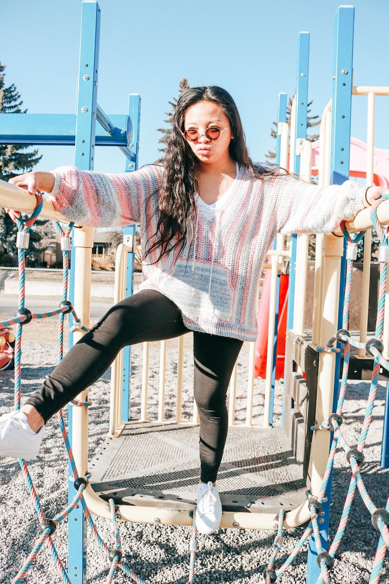 May include: A person wearing a pink and white striped sweater, black leggings, and white sneakers is standing on a playground structure. The structure has blue metal bars and ropes.