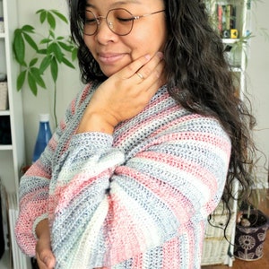 May include: A woman wearing a striped crochet sweater in shades of pink, blue, and white. The sweater has a loose fit and a relaxed style.