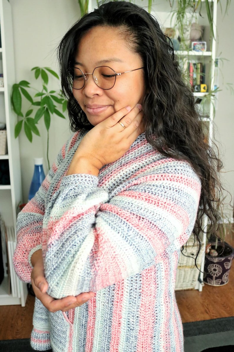 May include: A woman wearing a striped crochet sweater in shades of pink, blue, and white. The sweater has a loose fit and a relaxed style.