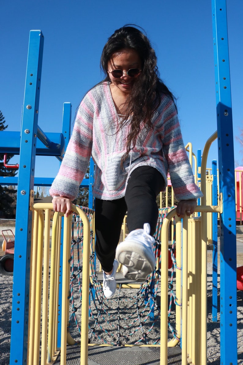 May include: A young person wearing a pink and white striped sweater and black pants is climbing on a yellow and blue playground structure. The person is wearing sunglasses and white sneakers.