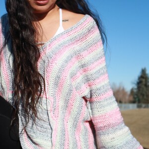 May include: A woman wearing a striped crochet sweater in shades of pink, gray, and white. The sweater has a loose fit and long sleeves.