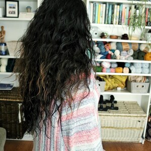 May include: A woman with long, curly, dark brown hair is wearing a striped crocheted sweater with pink, white, and gray stripes. The sweater has a loose fit and long sleeves.