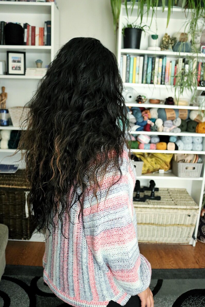 May include: A woman with long, curly, dark brown hair is wearing a striped crocheted sweater with pink, white, and gray stripes. The sweater has a loose fit and long sleeves.
