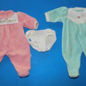 May include: A set of two long-sleeved footed sleepers and one pair of white briefs. One sleeper is pink with a white scalloped collar and floral embroidery, while the other is mint green with a white yoke featuring small red flower accents.