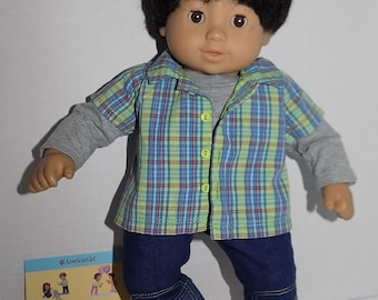 Puede incluir: Una muñeca vestida con una camisa a cuadros, una camisa de manga larga gris, pantalones vaqueros azules y zapatos rojos. La muñeca tiene el pelo negro y los ojos marrones. La muñeca sostiene un libro que dice "Bitty Twins Learn to Share".