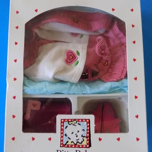 May include: A boxed Bitty Baby outfit set. The set includes a pink hat, jacket, and shoes, along with a white top with a rose design. The box is white with red hearts and the text "American Girl" and "Bitty Baby".