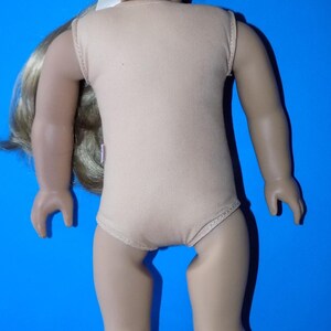 K&ouml;nnte beinhalten: Puppe mit blonden Z&ouml;pfen, tr&auml;gt einen beigefarbenen Body. Die Puppe hat einen hellen Teint und posiert vor blauem Hintergrund. Die Augen der Puppe sind geschlossen, und sie l&auml;chelt leicht.