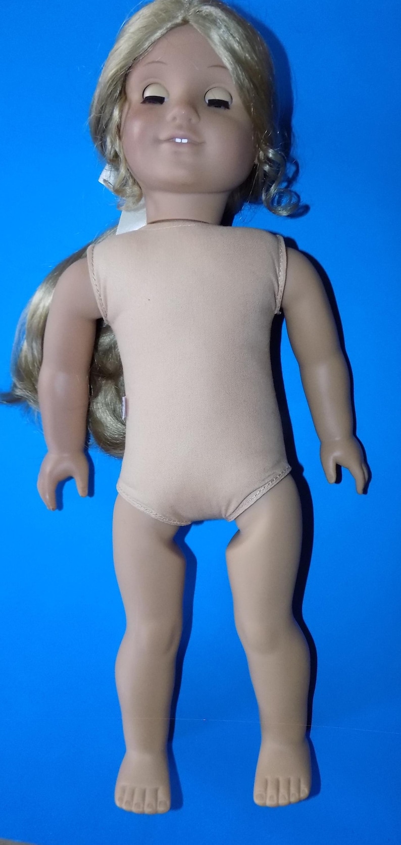 K&ouml;nnte beinhalten: Puppe mit blonden Z&ouml;pfen, tr&auml;gt einen beigefarbenen Body. Die Puppe hat einen hellen Teint und posiert vor blauem Hintergrund. Die Augen der Puppe sind geschlossen, und sie l&auml;chelt leicht.