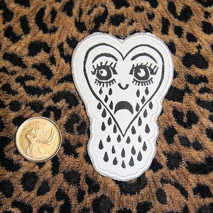 May include: White felt patch in the shape of a heart with a black outline. The heart has a sad face with large eyes and eyelashes, and is crying black teardrops.