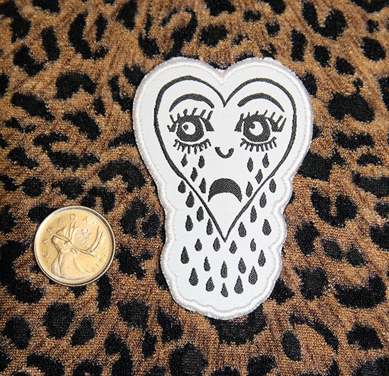 May include: White felt patch in the shape of a heart with a black outline. The heart has a sad face with large eyes and eyelashes, and is crying black teardrops.