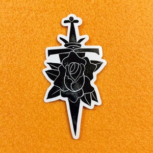 May include: Black and white sticker of a rose with a dagger through it.