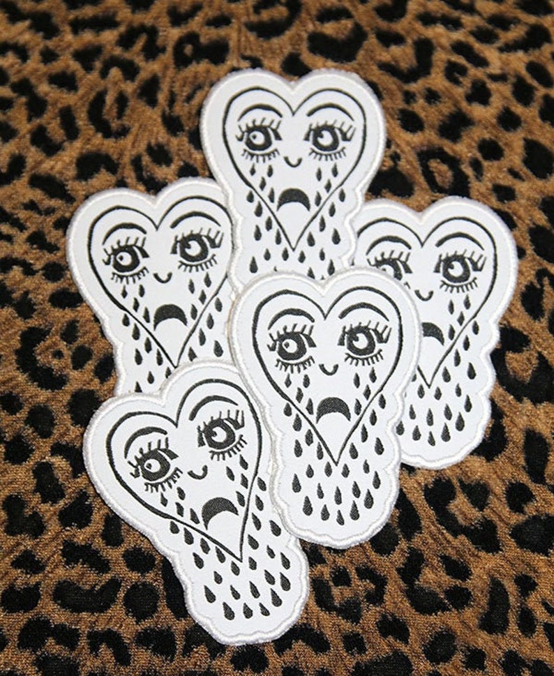 May include: Five white heart-shaped patches with black outlines and black tear drop designs. Each heart has a black face with large eyes and a sad expression.