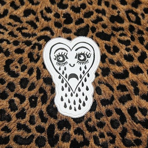 May include: A white heart-shaped patch with a black outline and a crying face. The heart has eyelashes, a frown, and black teardrops. The patch is on a brown and black leopard print fabric.