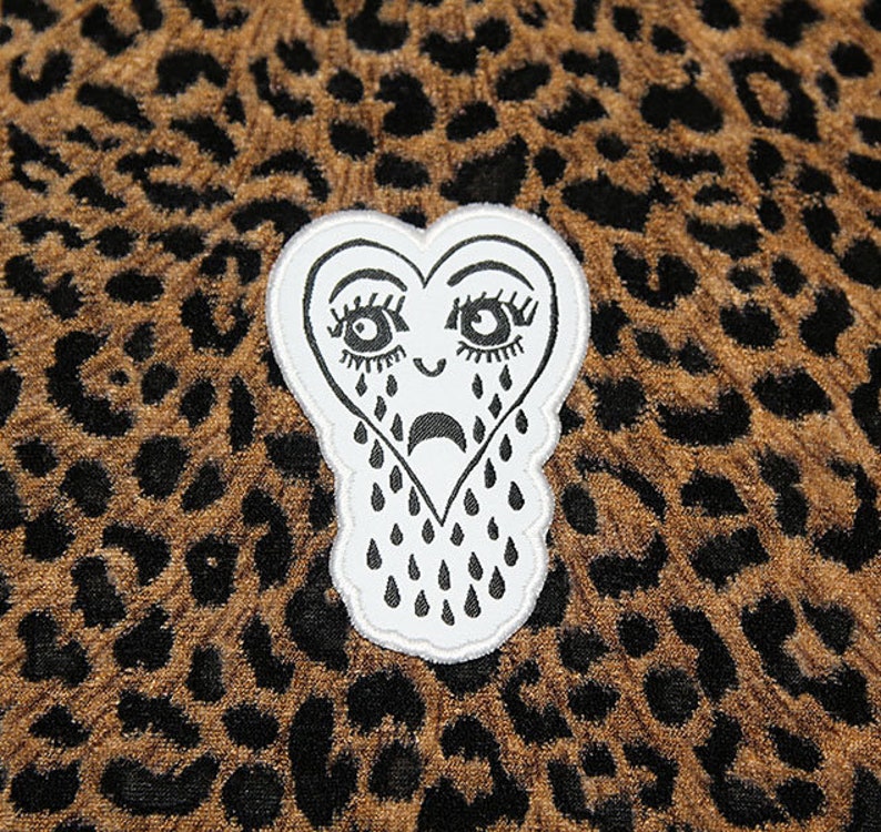 May include: A white heart-shaped patch with a black outline and a crying face. The heart has eyelashes, a frown, and black teardrops. The patch is on a brown and black leopard print fabric.