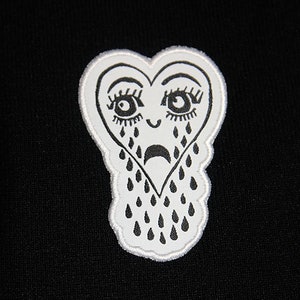 May include: White embroidered patch of a crying heart with eyelashes and teardrop shapes. The heart is outlined in white thread.