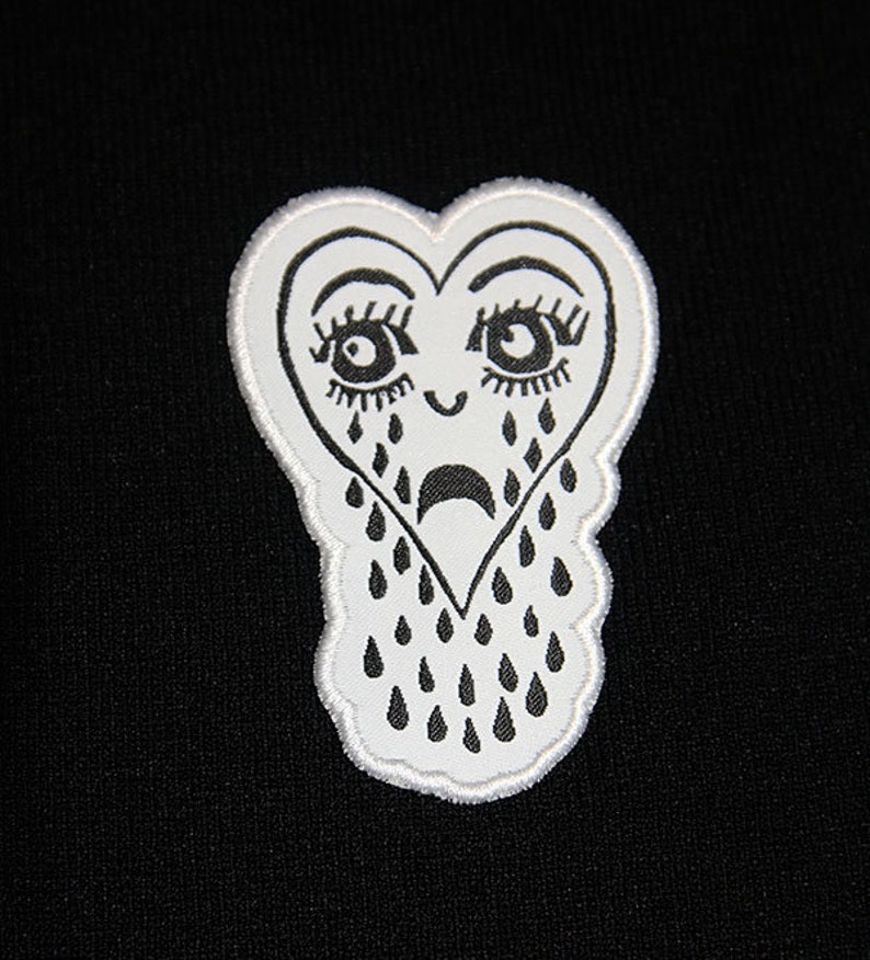 May include: White embroidered patch of a crying heart with eyelashes and teardrop shapes. The heart is outlined in white thread.