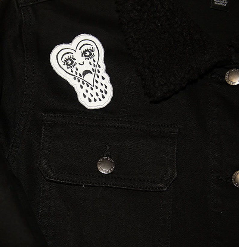 May include: Black denim jacket with a white heart-shaped patch featuring a crying face with teardrop designs.