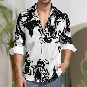 Puede incluir: Camisa blanca con un patrón abstracto negro. La camisa de manga larga tiene cuello y puños clásicos. El diseño negro se asemeja a remolinos de tinta sobre la tela blanca. La camisa está desabrochada en la parte superior.