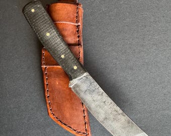 May include: A handcrafted knife with a dark handle and a weathered blade, resting in a brown leather sheath. The knife features a straight edge and a pointed tip. The sheath is stitched with a contrasting thread.