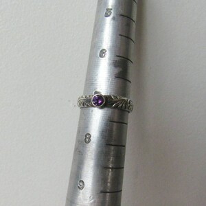 May include: A silver ring with a purple gemstone set in the center. The ring has a detailed design with a floral pattern.