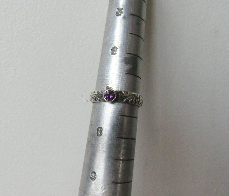 May include: A silver ring with a purple gemstone set in the center. The ring has a detailed design with a floral pattern.