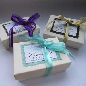 May include: Three white gift boxes with different colored ribbons. The boxes are tied with purple, gold, and turquoise ribbons. The boxes have a label that says "Xena Studio" with the text "silver, gold, & gemstones".