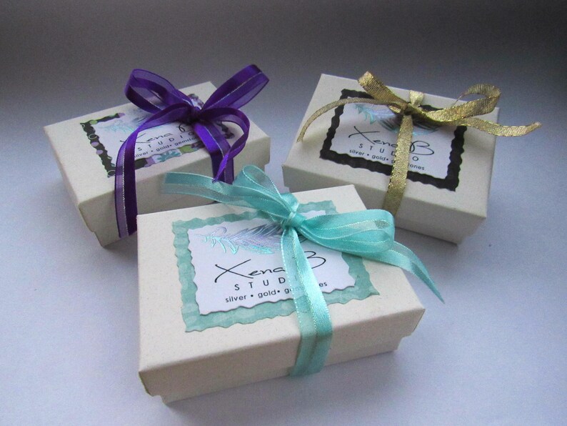 May include: Three white gift boxes with different colored ribbons. The boxes are tied with purple, gold, and turquoise ribbons. The boxes have a label that says "Xena Studio" with the text "silver, gold, & gemstones".