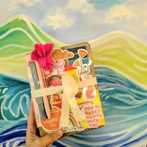 May include: A rectangular package wrapped in brown paper, decorated with various colorful stickers including text that reads "FORGIVEN", "beloved", and "GOD IS MADLY IN LOVE WITH YOU". It features a pink felt flower, a light pink ribbon, and two pens.
