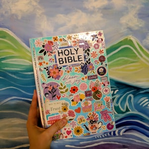 May include: A Holy Bible with a light blue cover decorated with numerous colorful floral and inspirational stickers. The text "HOLY BIBLE" is printed in black at the top center, with "bloom with joy" and "Trust" visible below.