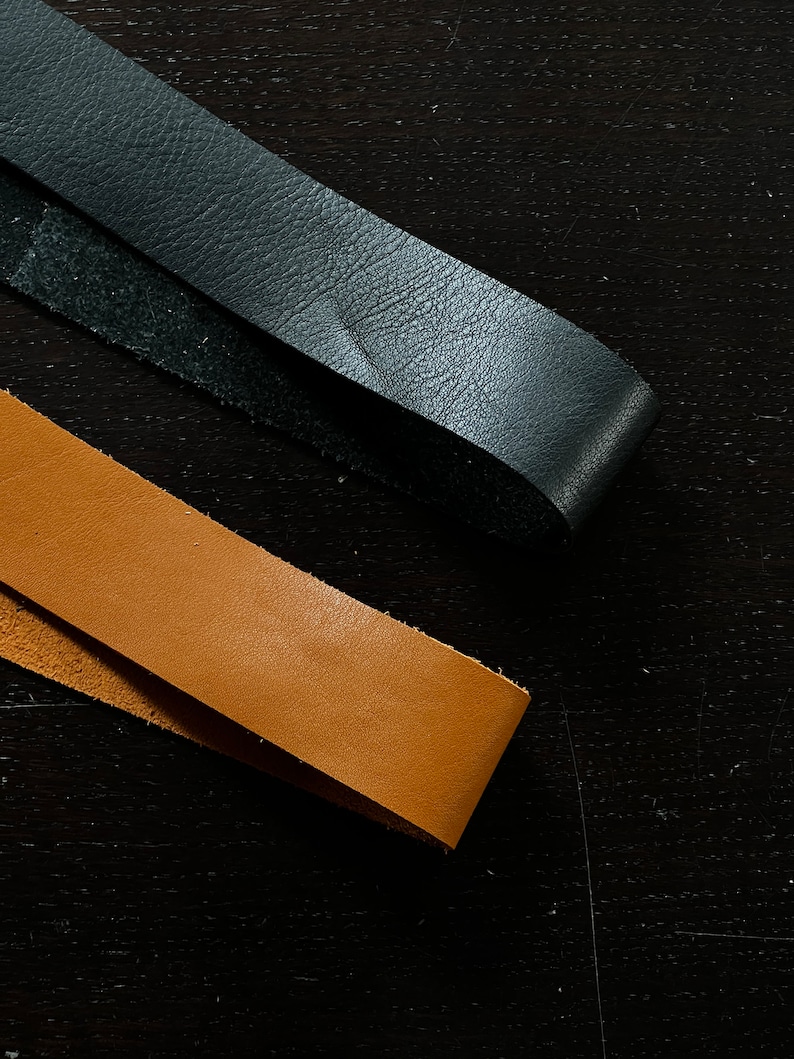 May include: Two leather straps, one black and one brown, are laid out on a dark wood surface.