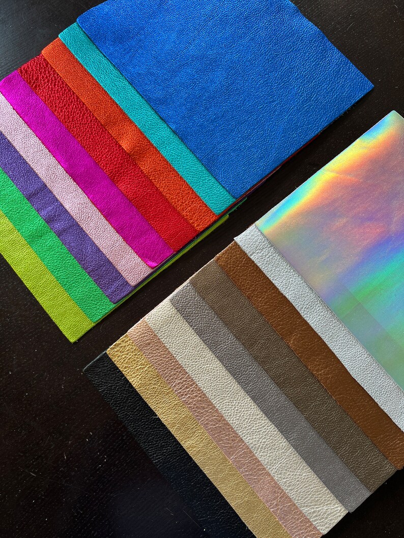 K&ouml;nnte beinhalten: Eine Sammlung von 20 verschiedenen Lederproben in verschiedenen Farben, darunter Blau, Gr&uuml;n, Rot, Orange, Lila, Pink, Braun, Schwarz, Wei&szlig; und Gold. Die Proben sind f&auml;cherf&ouml;rmig angeordnet, wobei die irisierende Probe oben liegt.