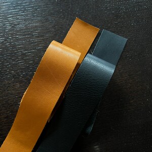 May include: Three leather strips, one black and two brown, are laid out on a dark wood surface.