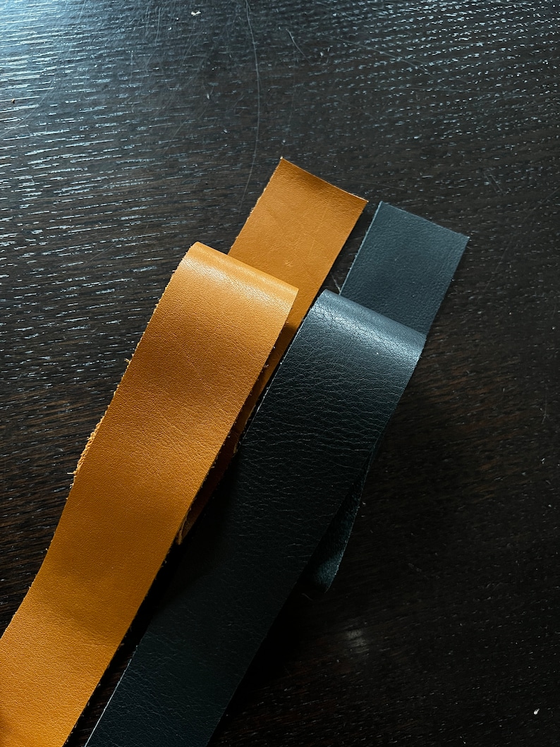 May include: Three leather strips, one black and two brown, are laid out on a dark wood surface.
