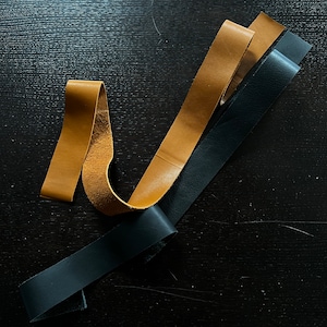May include: Three leather strips, one black and two brown, are arranged on a dark surface. The strips are different widths and lengths.