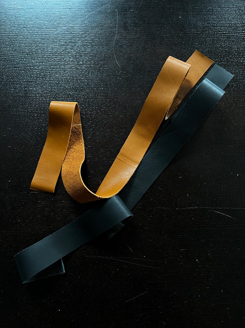 May include: Three leather strips, one black and two brown, are arranged on a dark surface. The strips are different widths and lengths.