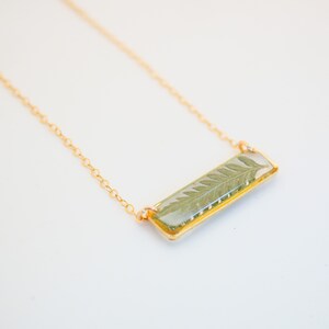 May include: A gold chain necklace with a rectangular pendant featuring a pressed fern leaf encased in clear resin.
