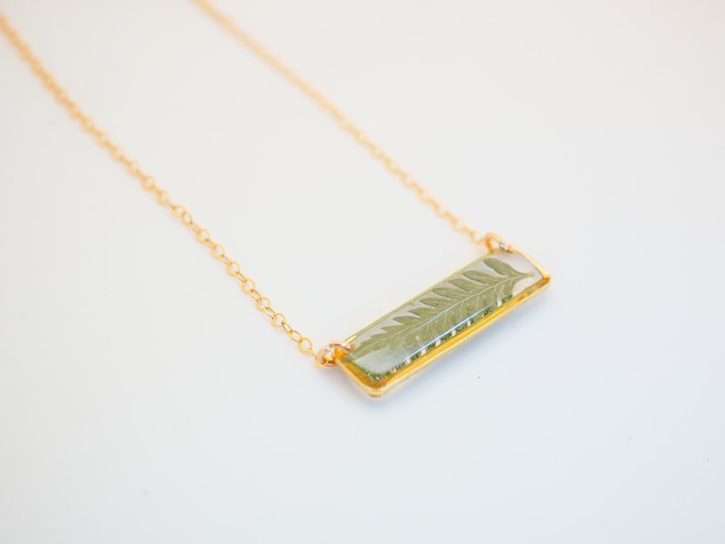 May include: A gold chain necklace with a rectangular pendant featuring a pressed fern leaf encased in clear resin.