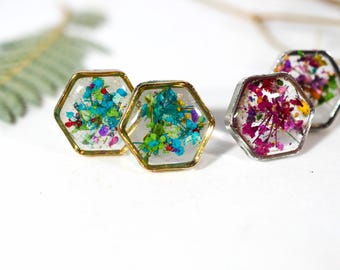 May include: Four hexagon-shaped resin rings, each containing dried flowers in various colours. Two rings have gold-coloured edges, and two have silver-coloured edges. The flowers are shades of blue, green, red, purple, and yellow. The rings are on a white background.