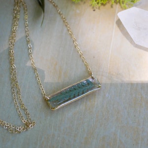 May include: A gold chain necklace with a rectangular pendant featuring a pressed fern leaf encased in clear resin.